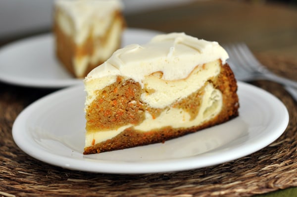 A piece of carrot cake cheesecake topped with white frosting, on a white plate. 