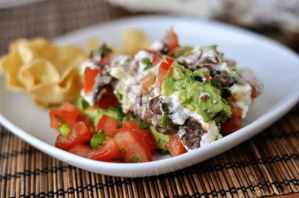 White plate with a big serving of 7-layer dip and tortilla chips on the side.