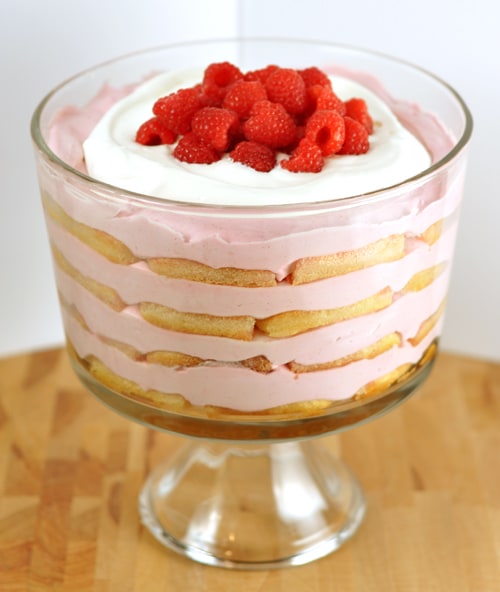 Clear trifle dish with layers of ladyfingers and pink berry layers and topped with whipped cream and fresh raspberries.