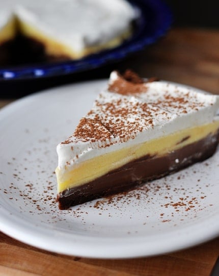 A slice of pie with a chocolate crust, pudding middle, and whipped cream top on a white plate.