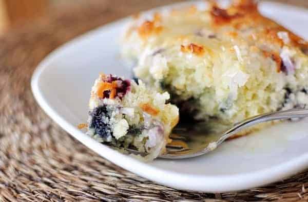 A piece of blueberry coconut cake on a white plate with a fork taking a bite out.