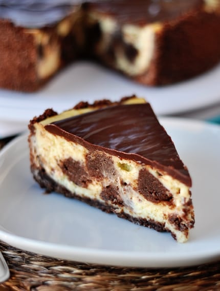 A piece of brownie-studded cheesecake topped with chocolate ganache on a white plate.