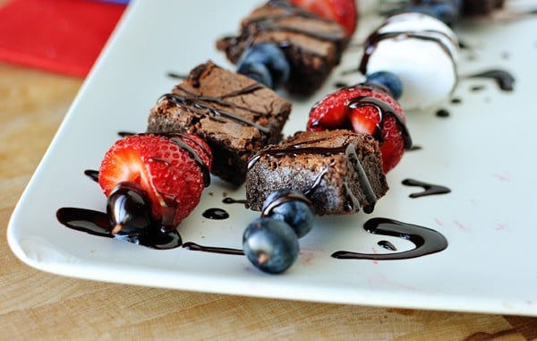 two brownie, marshmallow, and fruit kebabs drizzled with chocolate laying side by side on a white platter