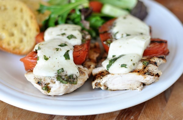 Grilled caprese chicken breasts in front of a salad and garlic bread on a white plate.