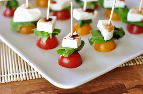 white platter with halved cherry tomatoes, a basil leaf, and a cube of fresh mozzarella skewered on toothpicks
