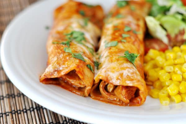 Two red sauce enchiladas on a white plate, with corn and a green salad on the side. 