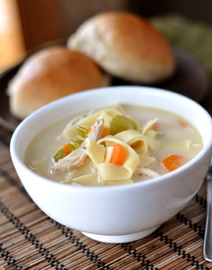 A white bowl full of chicken noodle soup with rolls in the background.