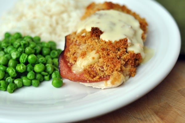 A chicken cordon bleu, with white sauce poured over the top and peas and potatoes on the side