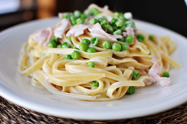White plate with creamy fettuccine, chopped ham, and green peas.