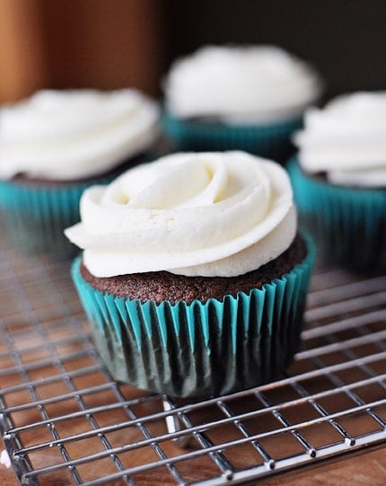 Frosting topped chocolate cupcakes in blue liners on a cooling rack.