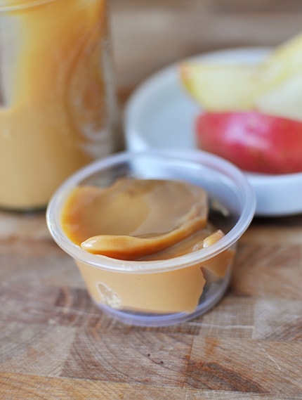 Small plastic container of dulce de leche with a mason jar of dulce and apple slices behind it.