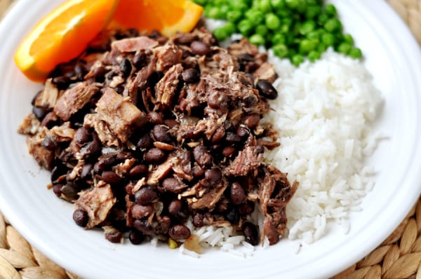 Top view of a plate of shredded pork and black beans, white rice, peas, and orange slices.