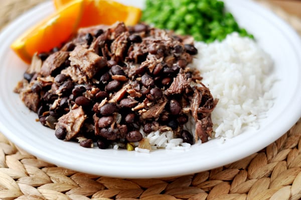 Shredded pork and black beans next to cooked white rice, peas, and orange slices on a white plate.