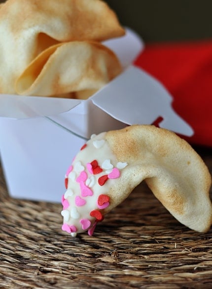 White container full of fortune cookies, with a half-dipped and sprinkled fortune cookie laying in front of the container.