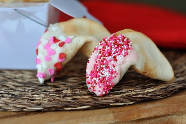 Two fortune cookies half dipped in Valentine's sprinkles in front of a white container.