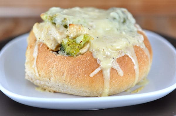 A bread bowl on a white plate with a chicken and broccoli mixture and melted cheese in the bowl.