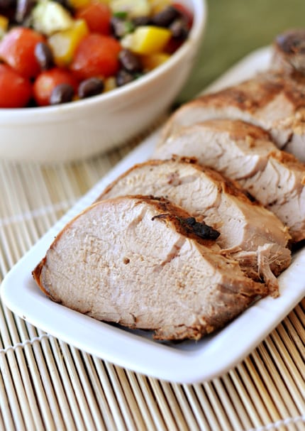 A white rectangular platter with sliced pork tenderloin next to a bowl of chopped veggies and black beans.