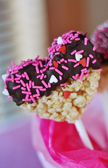 Heart-shaped, chocolate-dipped and sprinkle topped rice krispie treat on a white stick.