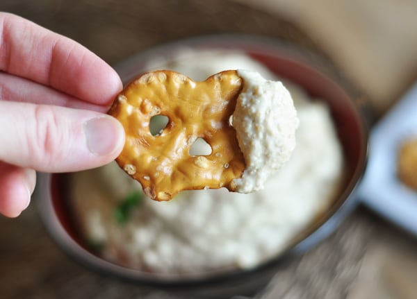 Someone holding a pretzel with hummus on one side over a bowl of hummus.