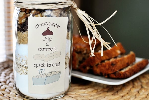 Layered dried ingredients in a mason jar for oatmeal chocolate chip quick bread, with the cooked bread sliced and laying on a platter in the background.