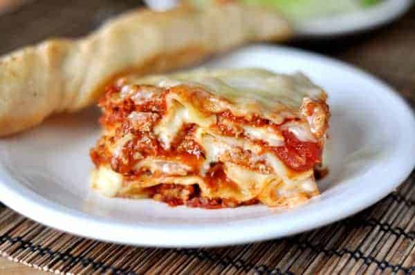 A big piece of meaty, saucy, cheesy lasagna sitting on a white plate with a breadstick laying next to it. 