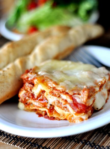 Piece of meat, cheese, and red sauce lasagna with breadsticks on a white plate.