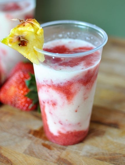 A coconut and strawberry swirled drink in a clear plastic cup with a wedge of pineapple on the top.