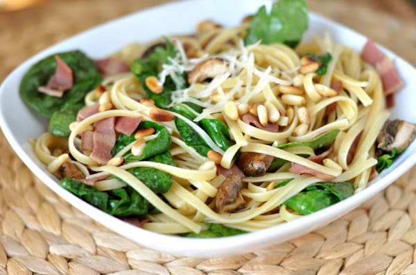 White bowl with cooked linguine, bacon, pine nuts and spinach.