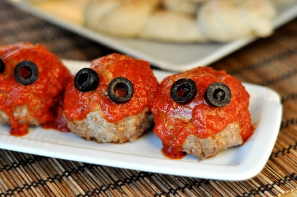 Three meatballs with red sauce and olive eyes lined up on a small rectangular white platter.