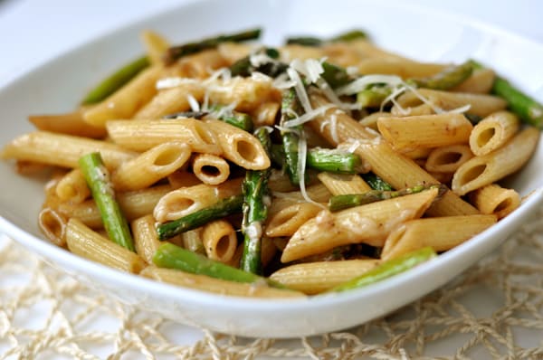 A white bowl of cooked penne pasta and pieces of asparagus.