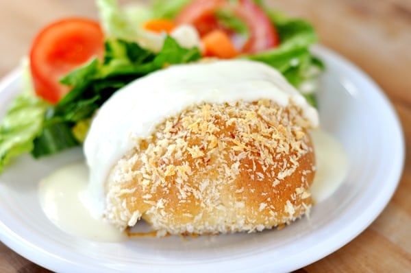 a white plate with chicken in a bread pillow, covered with a white sauce, and next to a green salad