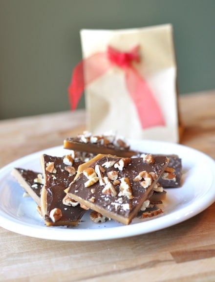 White plate of pieces of toffee buttercrunch with a paper sack and red ribbon behind it.