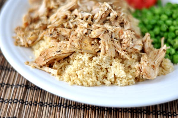 White plate with shredded pork atop a bed of cooked couscous and green peas on the side.