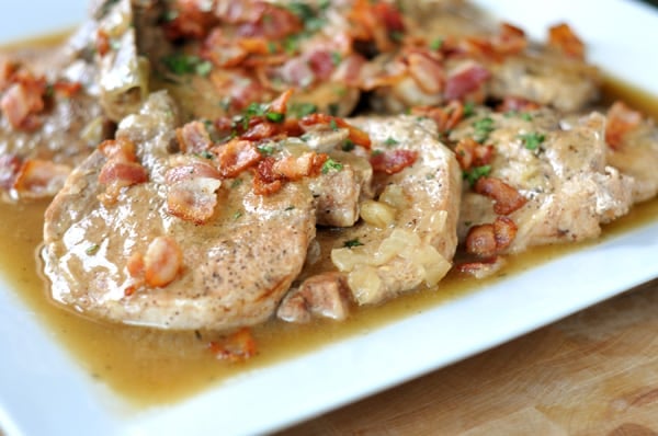 A white platter with cooked pork chops smothered in a brown sauce and chopped tomatoes.