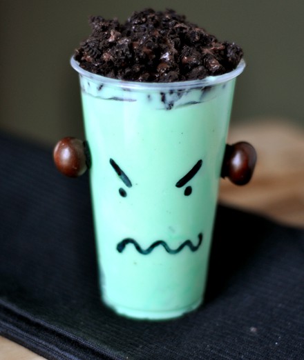 A plastic cup with green pudding, crushed Oreo's on top and a monster face drawn on the outside.