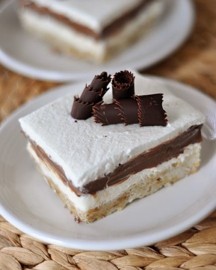 A piece of dessert on a white plate with a shortbread crust, pudding middle, and cool whip and chocolate curl topping.