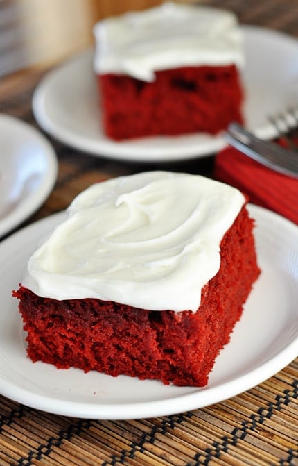 Two pieces of red velvet cake with buttercream frosting on white plates.