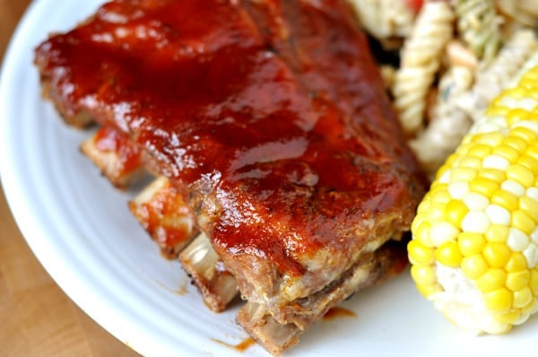A rack of bbq ribs on a white plate with corn on the cob and a pasta salad.