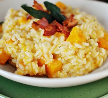 Cooked butternut squash risotto in a white bowl, with crumbled bacon and sage leaves on top.