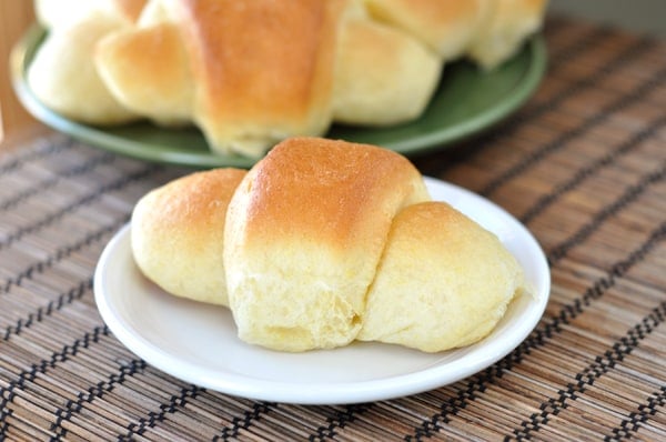 A white plate with a golden brown crescent roll with a green plate of more baked rolls behind it.