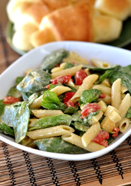 White dish with penne, spinach and tomato salad with a plate of crescent rolls behind it.