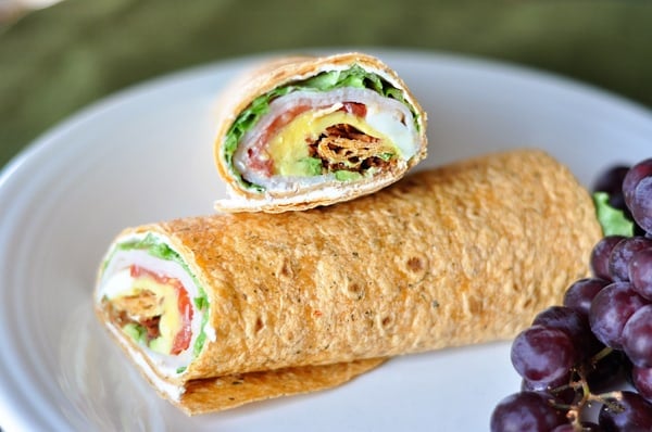 White plate with a turkey cobb wrap filled with veggies and turkey and cut in half next to purple grapes.