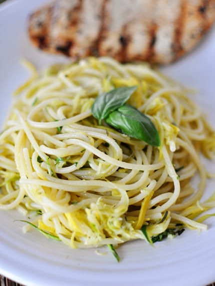 Cooked pasta with zucchini and yellow squash in it, and a few basil leaves on top and a grilled chicken breast behind it.