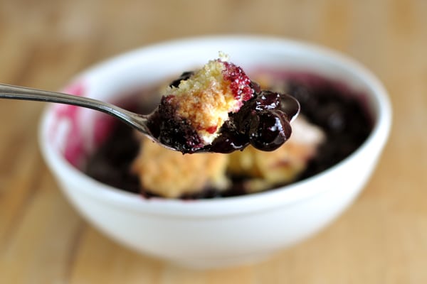 A white bowl of blueberry cobbler with a spoon in front holding a bite of it.