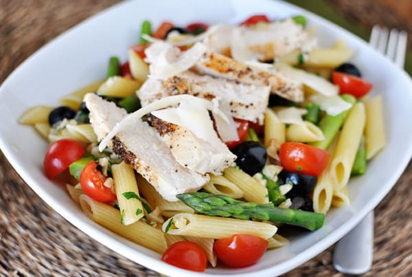 White bowl with cooked penne pasta, pieces of asparagus, cherry tomatoes, olives, and chicken slices.