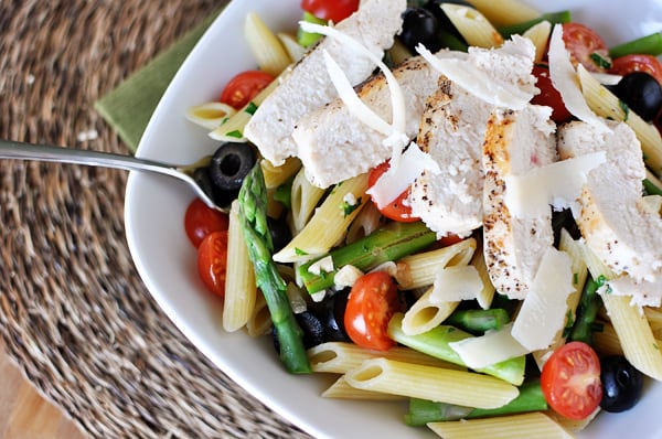 White bowl with cooked penne pasta, asparagus, tomatoes, olives, and chicken strips.