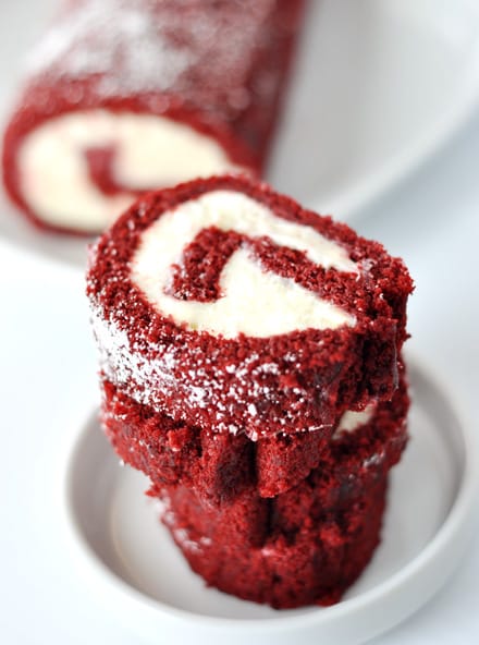 slices of a cream cheese filled red velvet spiraled cake roll stacked on a white plate, with the remaining roll behind it
