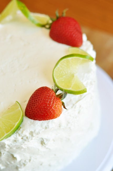A white platter with a frosted white cake topped with strawberries and ribbons of lime.