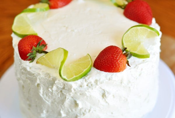 A white frosted cake on a white platter with ribbons of lime and full strawberries surrounding the top.