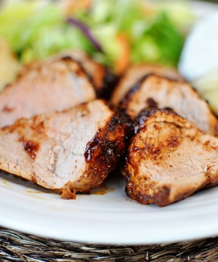 Slices of seasoned pork tenderloin on a white plate with a salad in the background.